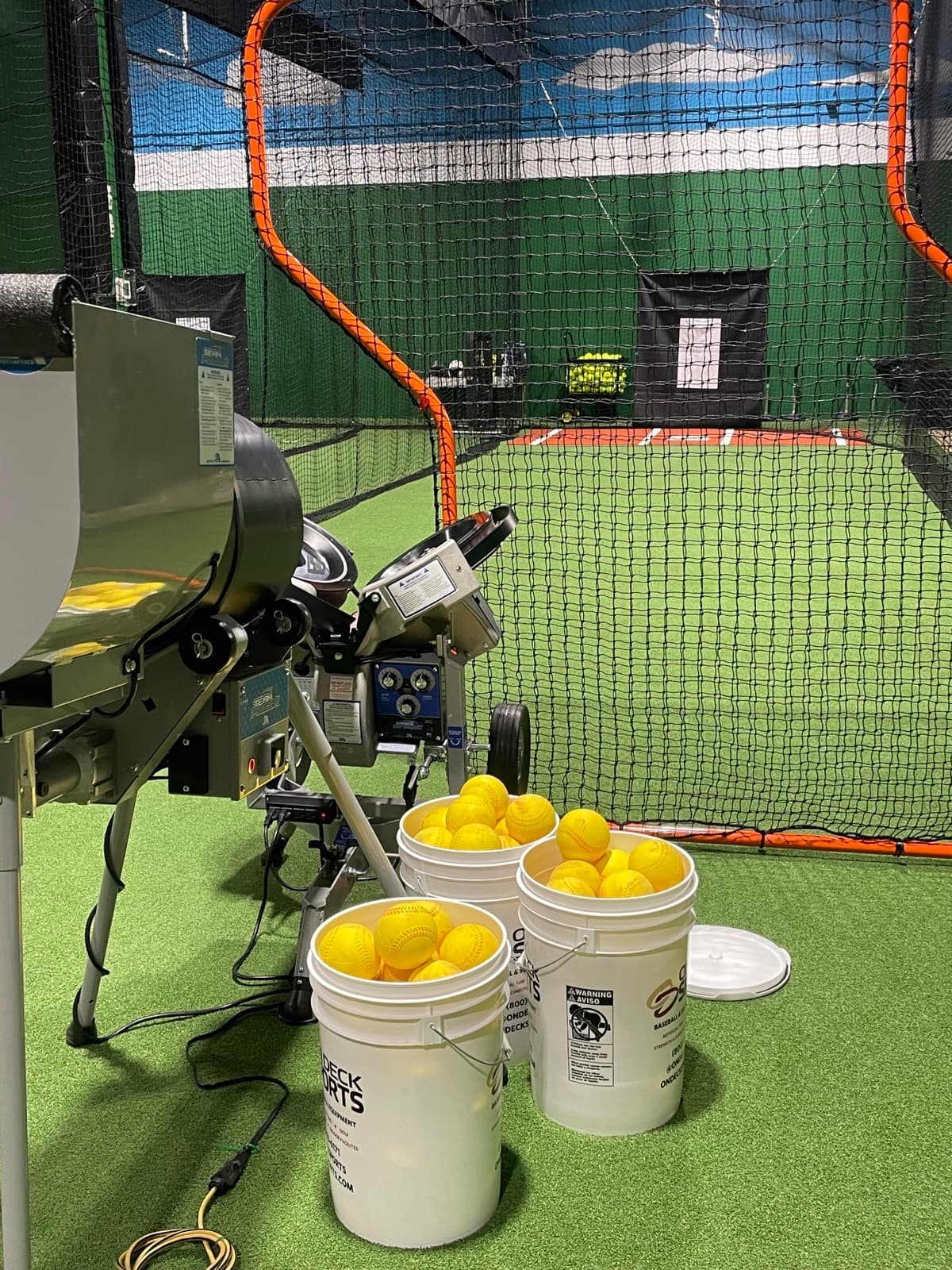 A pitching machine and buckets of yellow softballs on the turf inside a netted batting cage