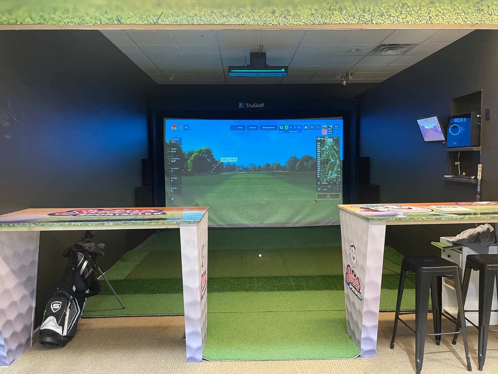 A TruGolf simulator bay with Swing Sportsplex branded high-top tables in front of the screen