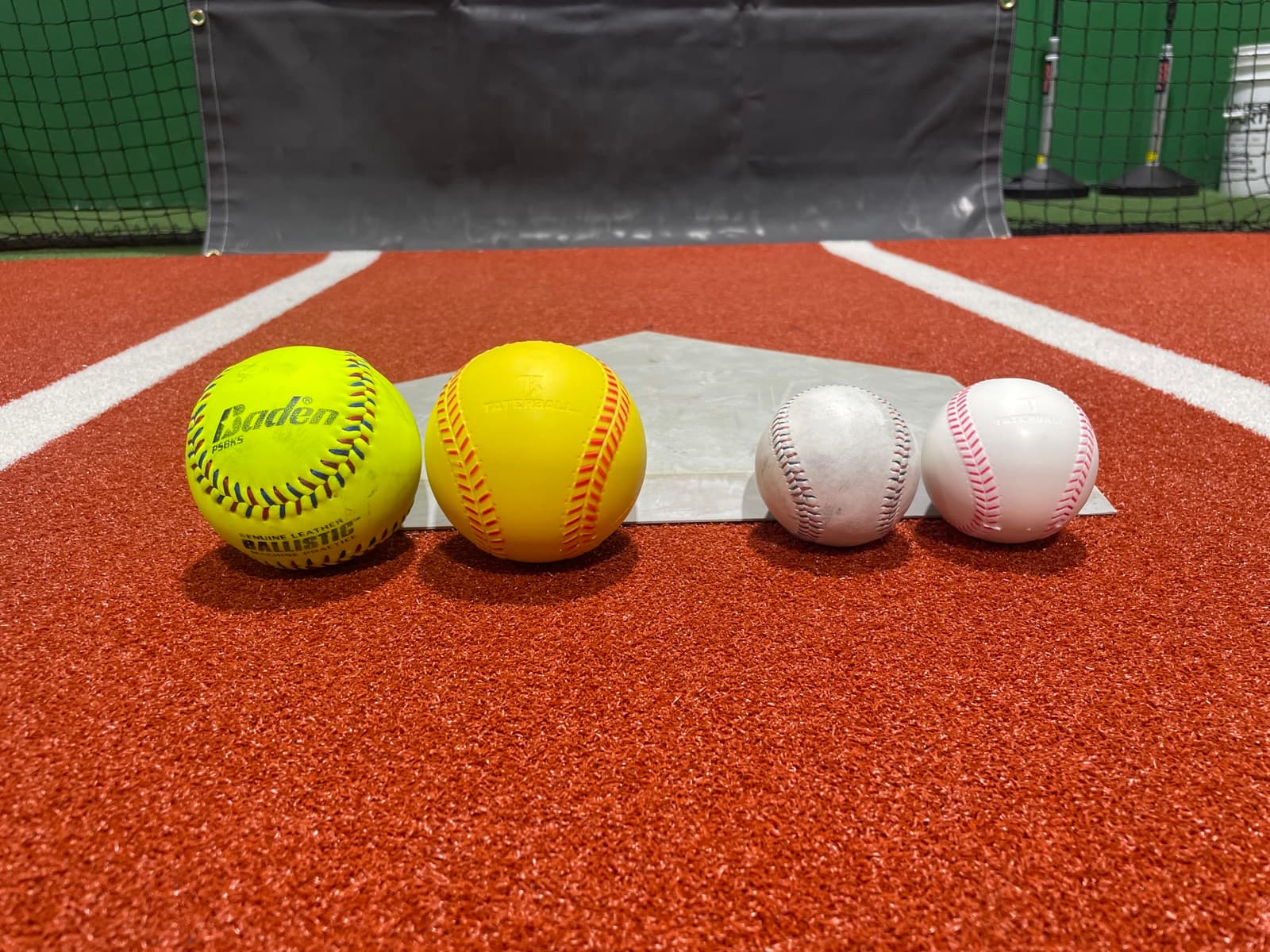 Two softballs and two baseballs lined up on home plate on the red turf