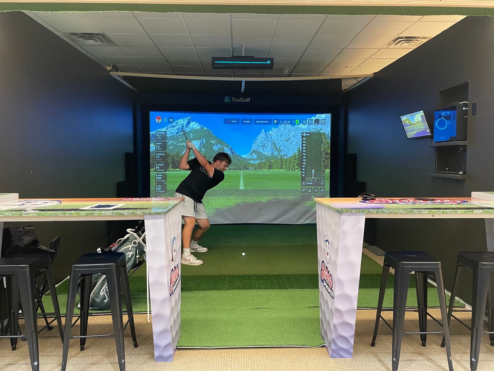 A golfer mid-swing on the TruGolf bay at Swing Sportsplex with branded high-tops and course on screen